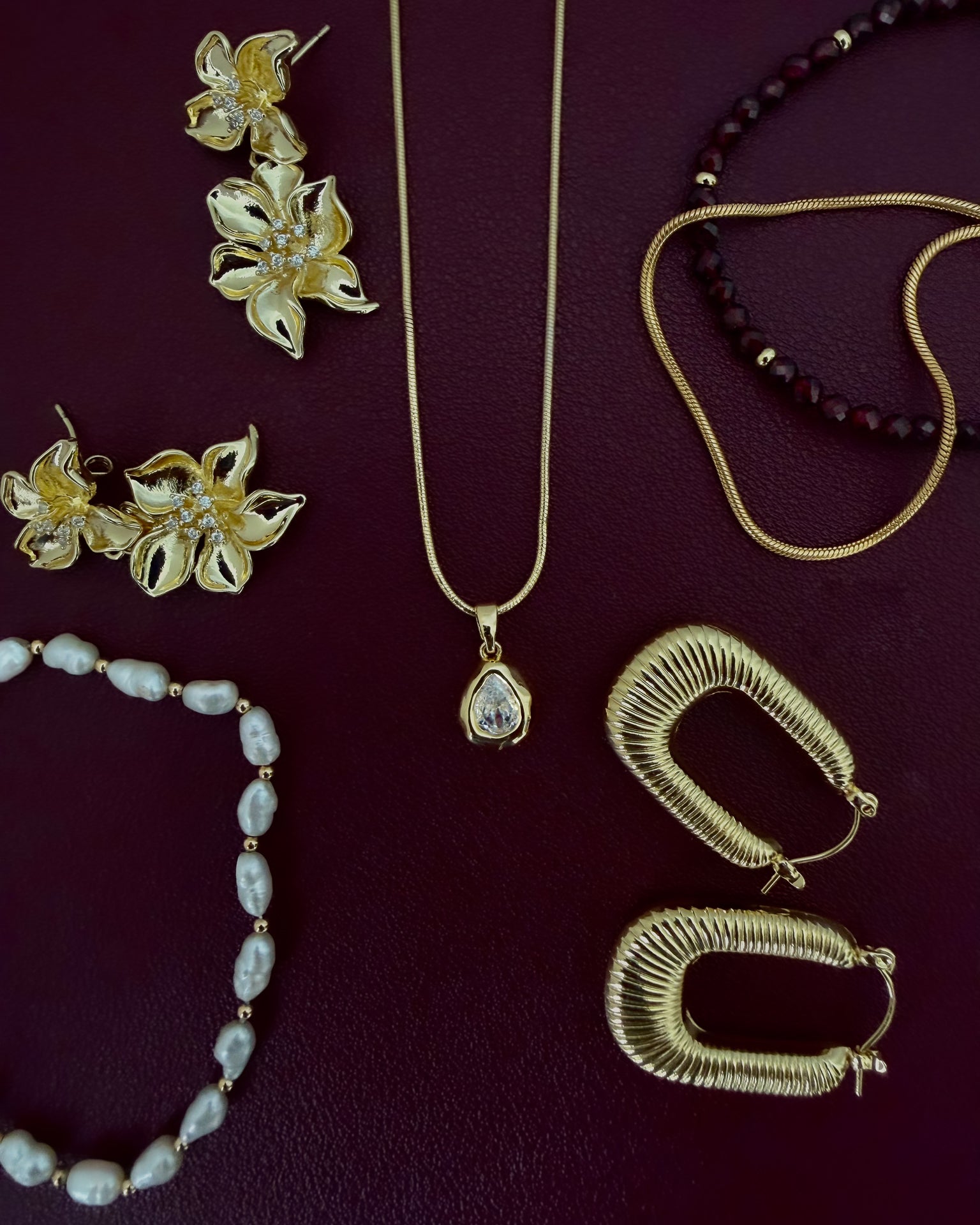 A flat-lay collection of gold and pearl jewelry on a dark burgundy background, including a gold pendant necklace, gold hoop earrings, gold flower earrings, a pearl bead bracelet, and a dark red beaded necklace.