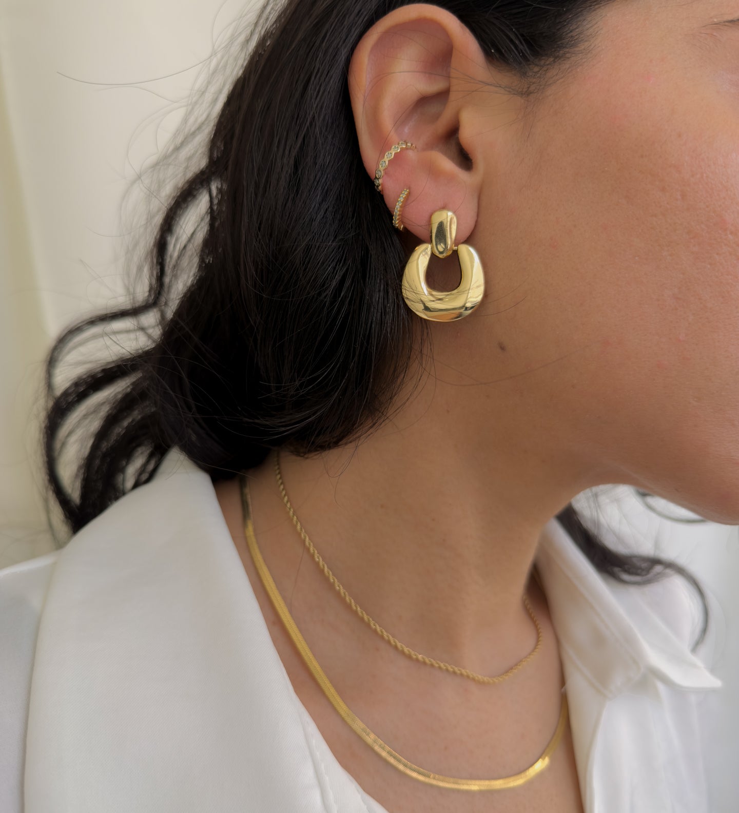 Close-up of a person wearing gold hoop earrings and necklaces against a neutral background