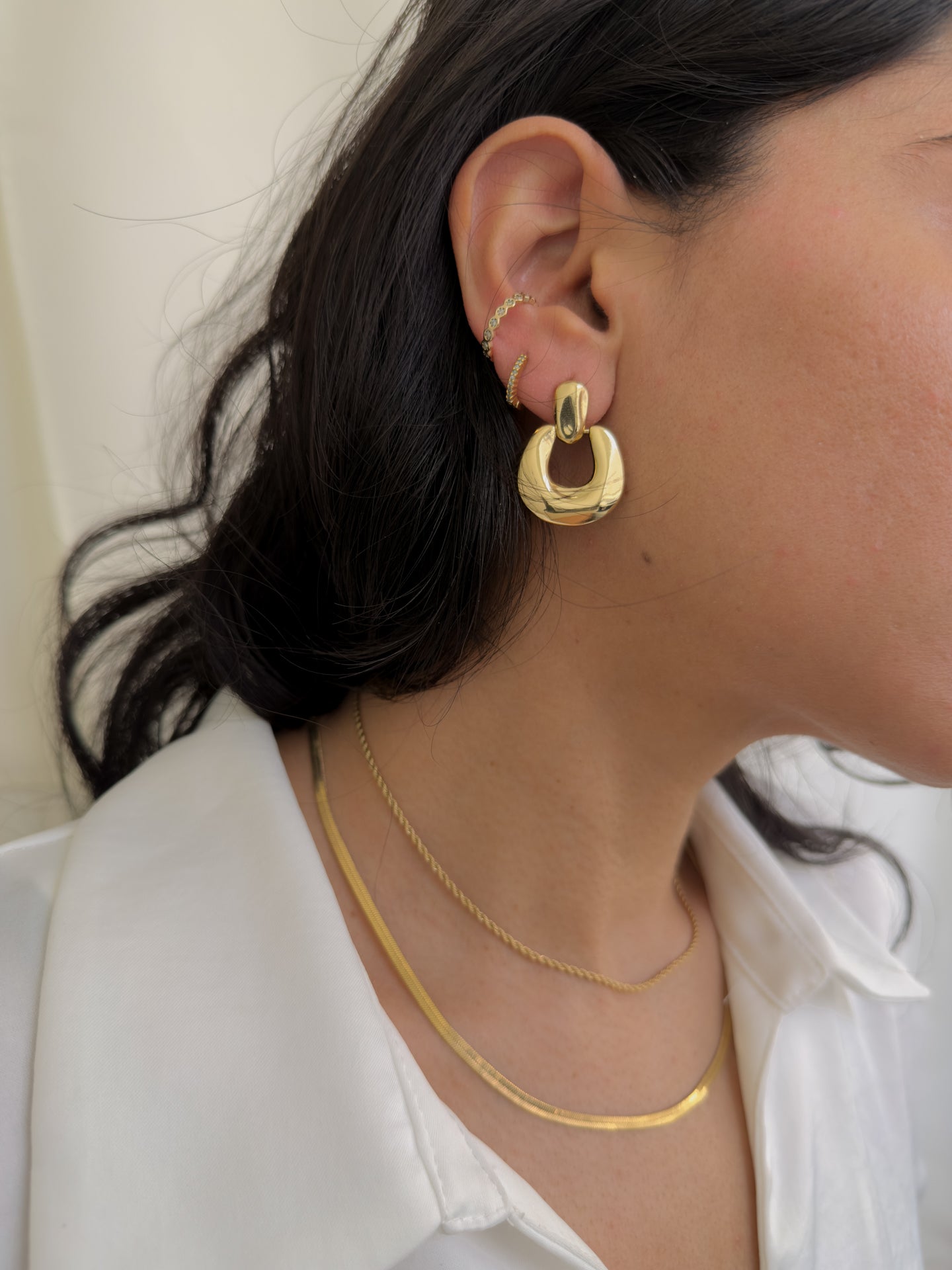 Close-up of a person wearing gold hoop earrings and necklaces against a neutral background