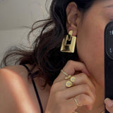 A close-up mirror selfie showing a person wearing a chunky, geometric gold earring, along with matching gold rings and a thin gold necklace. The person is holding a black smartphone to capture the reflection.