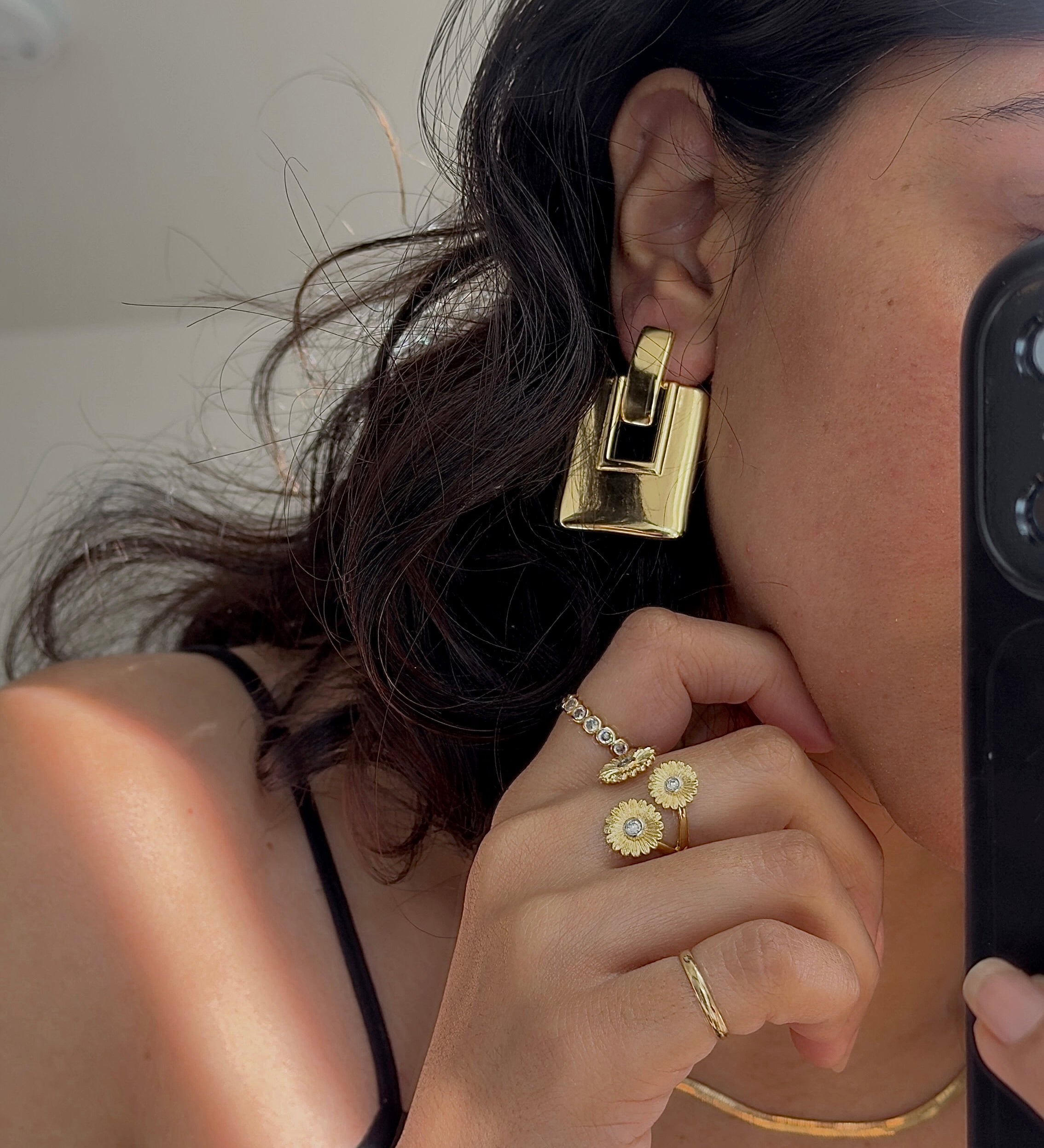 A close-up mirror selfie showing a person wearing a chunky, geometric gold earring, along with matching gold rings and a thin gold necklace. The person is holding a black smartphone to capture the reflection.