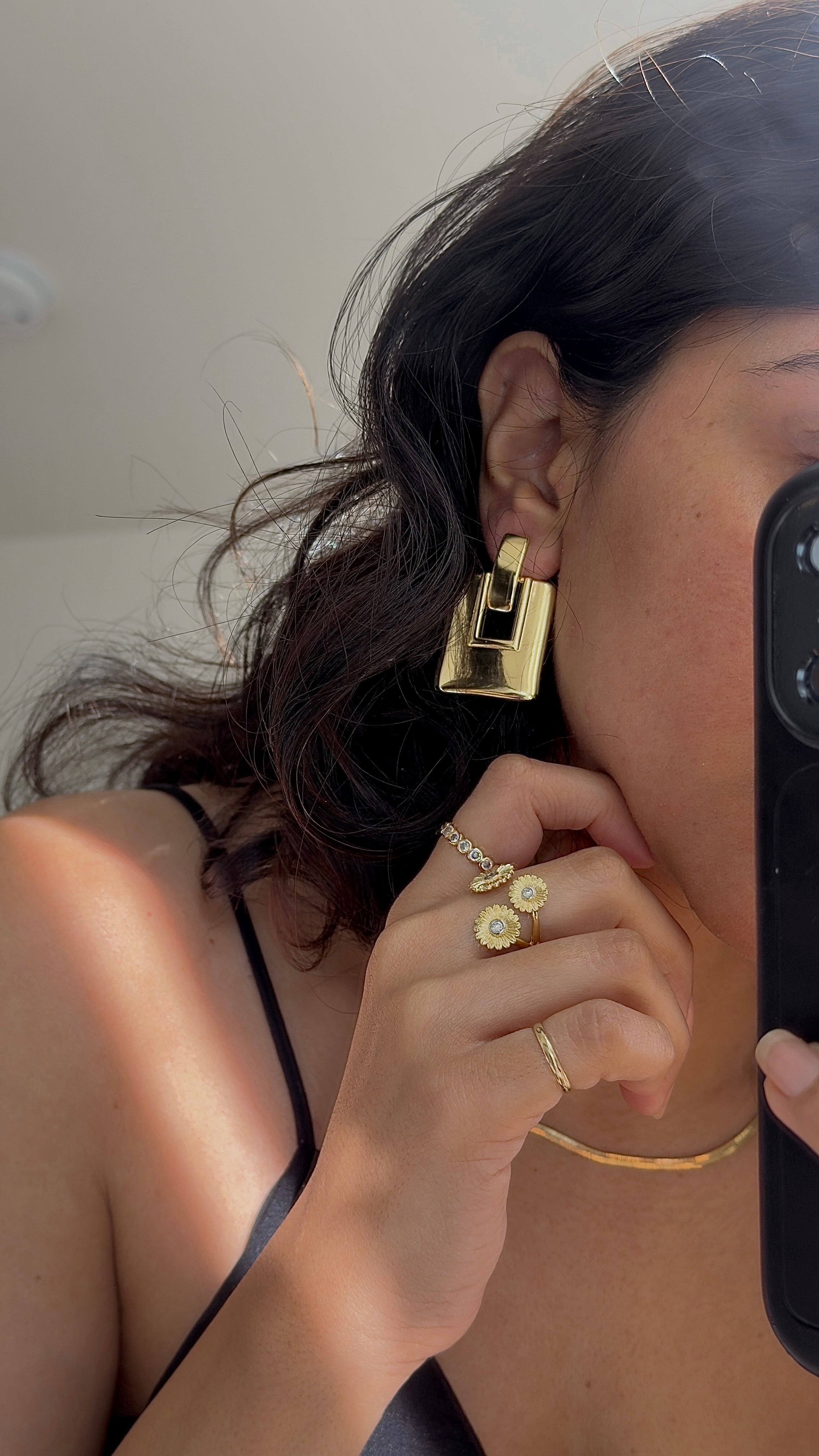 A close-up mirror selfie showing a person wearing a chunky, geometric gold earring, along with matching gold rings and a thin gold necklace. The person is holding a black smartphone to capture the reflection.